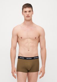 Barna pamut boxer alsónadrág fekete rugalmas derékpánttal, amelyen a "HUGO" márkajelzés látható. A modell semleges pózban áll egy fehér háttér előtt.