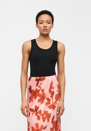 Femme aux cheveux bouclés courts portant un haut noir sans manches et une jupe rose avec des motifs floraux rouges, se tenant devant un fond blanc uni.