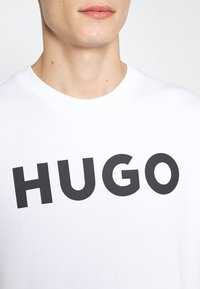 Camiseta de algodón blanca con cuello redondo que presenta letras negras en negrita que dicen "HUGO" en el pecho. Textura suave y ajuste casual.