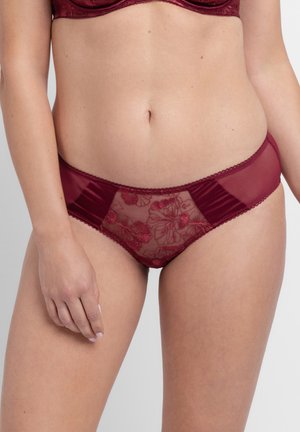 Nő bordó csipkés és hálós bikinistílusú fehérneműt és hozzáillő melltartót visel, világos egyszínű háttér előtt áll.