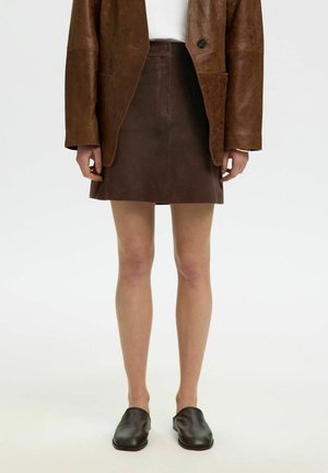 Personne portant un blazer en cuir marron, une mini-jupe en cuir marron foncé, un top blanc et des chaussures slip-on marron foncé, debout sur un fond blanc.