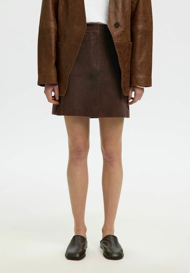 Personne portant un blazer en cuir marron, une mini-jupe en cuir marron foncé, un top blanc et des chaussures slip-on marron foncé, debout sur un fond blanc.