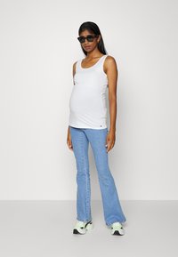 Top de alças branco com decote arredondado, combinado com jeans claros de boca de sino e ténis pretos com detalhes em verde. Tecido suave, design ajustado.