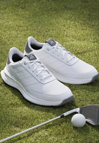 Scarpe da golf bianche con accenti grigi, materiale sintetico, superficie testurizzata e lacci. Posizionate sull'erba con un bastone da golf e una palla nelle vicinanze.
