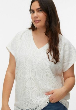 Kobieta o długich ciemnych włosach, ubrana w białą bluzkę z krótkim rękawem i wzorem oraz niebieskie dżinsy, pozująca z jedną ręką w kieszeni.