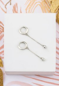 Silver hoopsmycken med kedja och små bollpendlar, visade på en vit fyrkantig ställning mot en mönstrad bakgrund.
