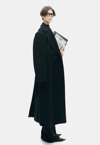 Lang zwarte jas met brede revers, enkele knoopsluiting en verlaagde schouders. Slanke zwarte coltrui en zwart puntige laarzen. Houdt documenten vast.