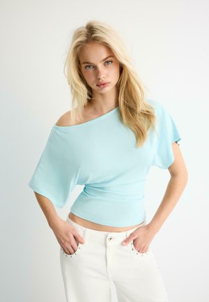 Giovane donna con lunghi capelli biondi che indossa una maglia azzurra scollata sulle spalle e pantaloni bianchi, in posa con le mani nelle tasche su uno sfondo bianco.