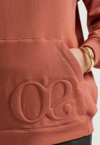Rustfarbenes Sweatshirt mit strukturiertem Finish, ausgestattet mit einer großen bestickten "03" auf der Vorderseite und einer Fronttasche. Glattes Material.
