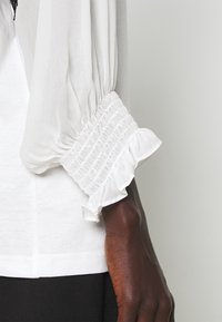 Blusa blanca con mangas transparentes y abullonadas, con puños fruncidos y volantes, y una textura suave y ligera.