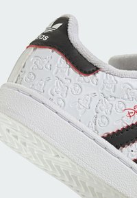 Baskets blanches avec personnages de dessin animé en relief, accents noirs et rouges, semelle en caoutchouc texturée, et bande noire sur le côté. Dotées d'un col rembourré.