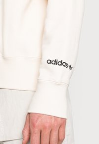 Felpa color crema con polsini a costine. Logo adidas nero sulla manica sinistra. Il materiale sembra morbido e con una texture.