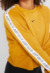 Bluza Nike w musztardowym kolorze, z białymi paskami i napisem "JUST DO IT". Ściśnięte mankiety i dół, miękka tekstura, luźny fason.
