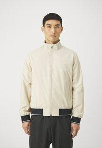 Giacca zip-up beige chiaro con colletto alto, polsini elasticizzati e dettagli neri sull'orlo e sui polsini, realizzata in tessuto liscio.