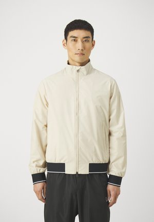 Lichtbeige ritsjack met een hoge kraag, elastische manchetten en zwarte accenten op de zoom en manchetten, gemaakt van een gladde stof.