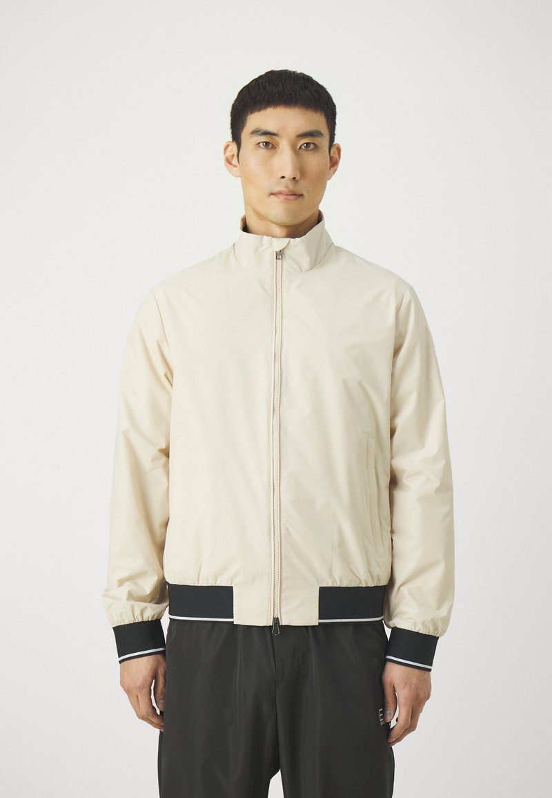 Giacca zip-up beige chiaro con colletto alto, polsini elasticizzati e dettagli neri sull'orlo e sui polsini, realizzata in tessuto liscio.