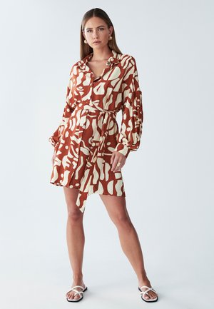 Sukienka zapinana na guziki w kolorze rdzy z kremowymi abstrakcyjnymi wzorami. Posiada długie rękawy, wiązanie w talii oraz asymetryczny dół, który jest krótszy z jednej strony.