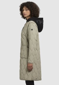 Chaquetón beige acolchado con capucha negra, que cuenta con un logotipo, bolsillos laterales y un diseño a la altura de la rodilla. Textura suave y silueta moderna.