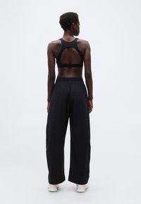 Άτομο με την πλάτη γυρισμένη, φοράει μαύρο αθλητικό σουτιέν, άνετο μαύρο παντελόνι, λευκά αθλητικά παπούτσια, στέκεται μπροστά σε ένα λευκό σκηνικό.