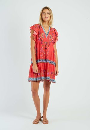 Woman standing wearing a red floral dress with ruffled sleeves and blue patterned borders, paired with tan heeled sandals on a white background.