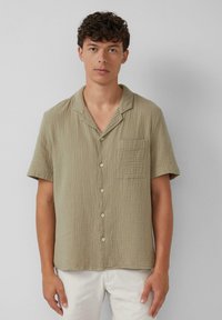 Kurzärmliges Button-up-Hemd in Olivgrün, aus zerknittertem Stoff, mit einer Brusttasche und einem lockeren Schnitt. Einfache weiße Knöpfe.