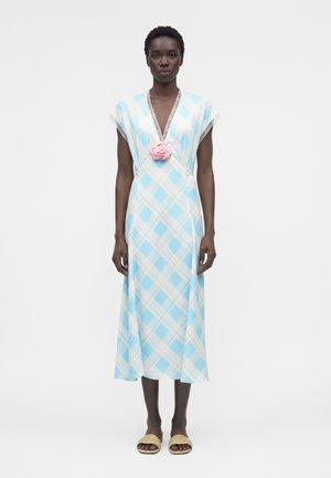 Kobieta ubrana w niebiesko-białą sukienkę midi w kratę z koronkowym wykończeniem, głębokim dekoltem w serek, różową broszką w kształcie rozety oraz beżowymi sandałami wsuwanymi, stojąca na białym tle.