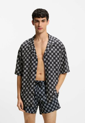 Giovane uomo con camicia aperta e pantaloncini abbinati con il motivo a logo "HUGO" in bianco e nero, in piedi con una mano in tasca su sfondo bianco.