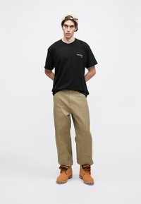 Tricou negru din bumbac cu un logo mic, pantaloni bej cu picioare largi și cizme bej cu șireturi. Modelul stă cu mâinile în spate pe un fundal simplu.