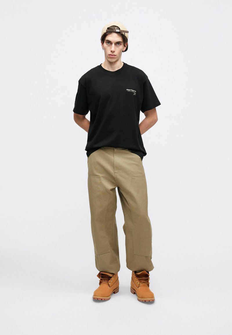 Svart bomullströja med litet logotyp, beige vida byxor och tanfärgade kängor med snören. Modellen står med händerna bakom ryggen mot en enkel bakgrund.