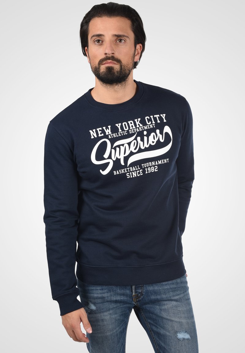 Marineblå sweater med rund hals; med hvide trykte bogstaver der siger "New York City Superior Basketball Tournament Siden 1982."