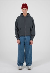 Grauer Kapuzenpullover mit Reißverschluss und Fronttaschen, getragen über hellblauen, weit geschnittenen Jeans. Accessoires: rote, strukturierte Mütze und silberne Sneaker.