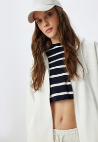 Felpa bianca con zip sopra un crop top a righe blu navy e bianche, con un cappello chiaro e capelli texturizzati. Sfondo minimalista, toni neutri.