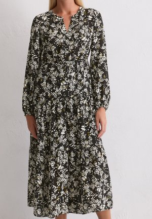 Vrouw gekleed in een lange zwarte jurk met witte en gele bloemenprint, lange mouwen en een V-hals, staand tegen een effen muur.