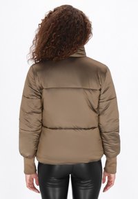 Brązowa kurtka puchowa z wysokim kołnierzem i prążkowanymi mankietami, z pikowanymi sekcjami i gładkim, błyszczącym wykończeniem. Czarne legginsy widoczne poniżej.
