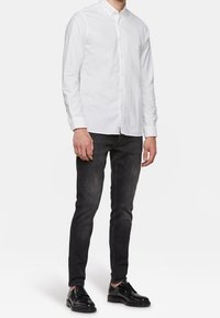 Man står klädd i en vit långärmad skjorta med knappar, figursydda svarta jeans och svarta läderskor.