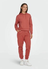 Vrouw staat en glimlacht, gekleed in een stofroze hoodie en bijpassende joggingsbroek, met witte sneakers en sokken.