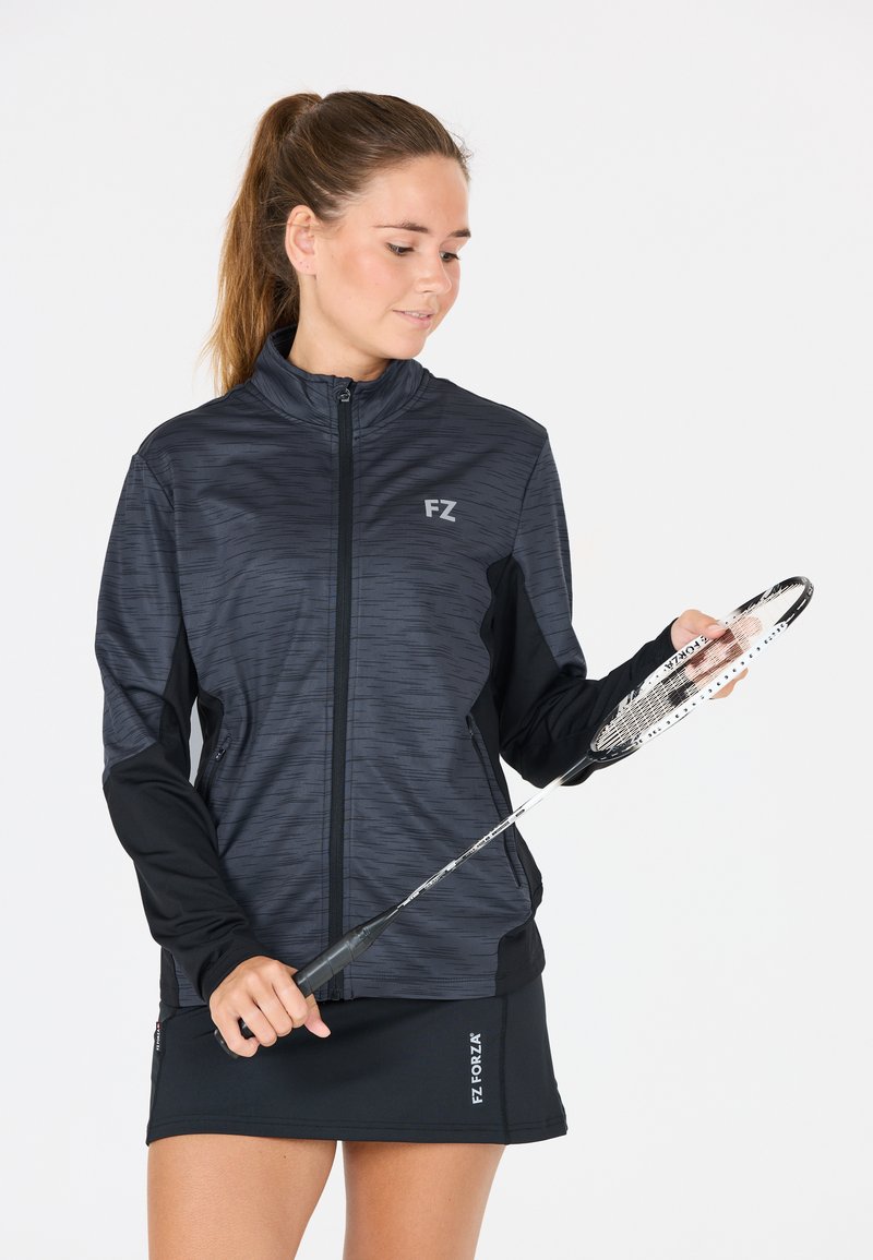 Kvinna i svart sportjacka och kjol håller en badmintonracket och ser ner på den mot en enfärgad vit bakgrund.