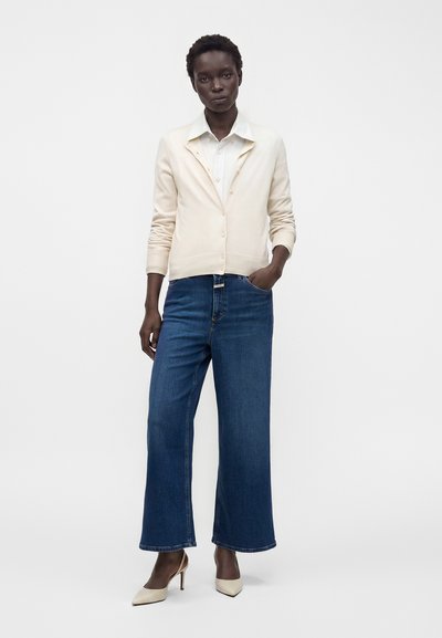 Femme debout portant un cardigan crème boutonné, une chemise blanche à col, un jean bleu jambes larges et des chaussures crème à talons hauts sur fond uni.