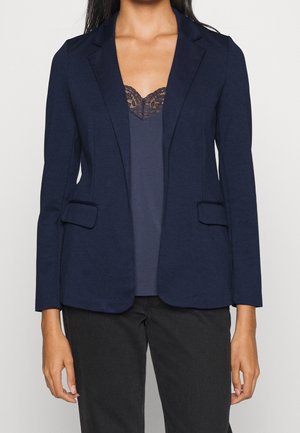 Vrouw draagt een marineblauwe blazer over een donker kanten topje en zwarte broek, getoond van schouders tot heupen.