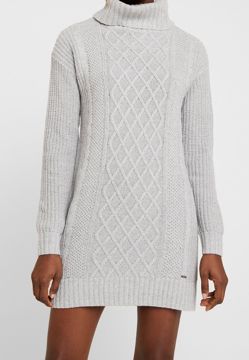 Robe-pull à col roulé en maille torsadée gris clair à manches longues portée par une personne à la peau foncée sur un fond uni.