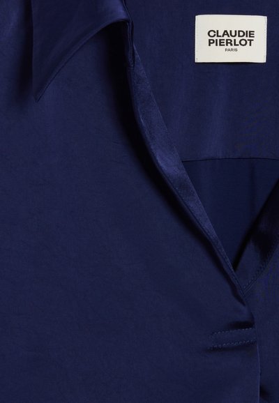 Blazer in tessuto blu navy con una texture liscia, caratterizzato da un colletto a punta e una tasca cucita. Etichetta con la scritta "CLAUDIE PIERLOT PARIS" visibile.