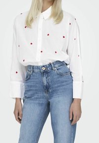 Chemise blanche à boutons avec des motifs de cœurs rouges, à manches longues et poignets. Associée à un jean en denim bleu taille haute. Design et coupe simples.