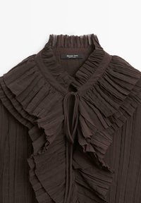 Blouse plissée brun foncé avec un col à volants haut et fermeture à nouer. Fabriquée dans un tissu léger, présentant des couches texturées et un fin détail.