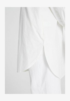 Valge puuvillane särk kõvera servaga, mis on valmistatud kergekangast ja tekstuurilisest materjalist, külgmiste lõigetega mugavaks liikumiseks.