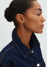 Giacca in denim scuro con un ampio colletto, caratterizzata da cuciture arancioni a contrasto e dettagli in metallo argentato, mostrata in profilo su uno sfondo neutro.