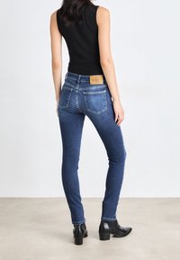 Jeans ajustados en un denim azul oscuro con una cintura de media altura y una etiqueta de cuero en el bolsillo trasero. Combinados con botas negras hasta el tobillo.