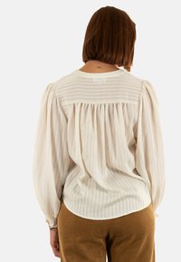 Blusa blanca con rayas texturizadas, corte suelto y mangas abullonadas. Tejido suave con un detalle en la parte posterior fruncido, combinada con pantalones de pana marrón.