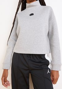 Šedá zkrácená mikina s vysokým límcem a černým logem na hrudi, doplněná o černé teplákové kalhoty se bočním bílým pruhem.