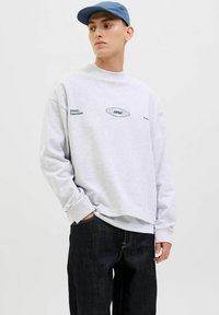 Sudadera gris claro de manga larga, con puños acanalados y texto impreso; combinada con jeans vaqueros oscuros; diseño casual, textura suave.