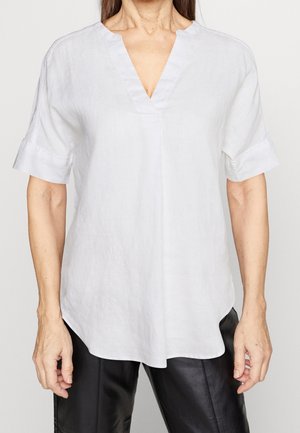 Frau trägt eine lockere weiße V-Ausschnitt-Kurzarmbluse mit umgeschlagenen Manschetten und schwarze Lederhose, gezeigt vom Hals bis zur Mitte des Oberschenkels.